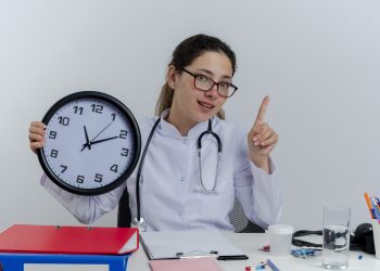 impressed young female doctor wearing medical robe and stethoscope and glasses sitting at desk with medical tools looking at camera holding clock raising finger isolated on white background