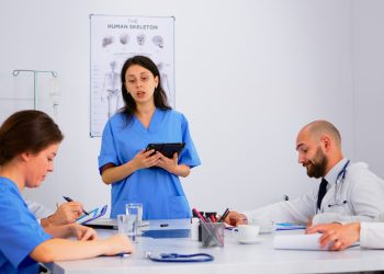 Medical people meeting and planning with shareholders in hospital office sitting at desk. Doctors and nurses brainstorm ideas together, physicians diagnosis and presentation data using tablet