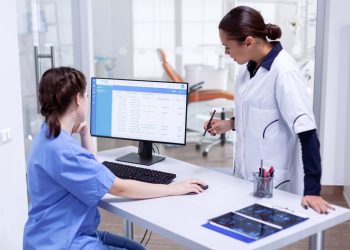 Stomatolog and nurse in tooth clinic checking patient appointment looking at computer monitor. Stomatology assistant and teeth doctor discussing in reception of dental office