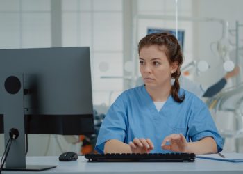 Stomatological assistant typing on computer keyboard sitting at oral clinic desk. Nurse using technology while professional orthodontist consulting patient with equipment in background